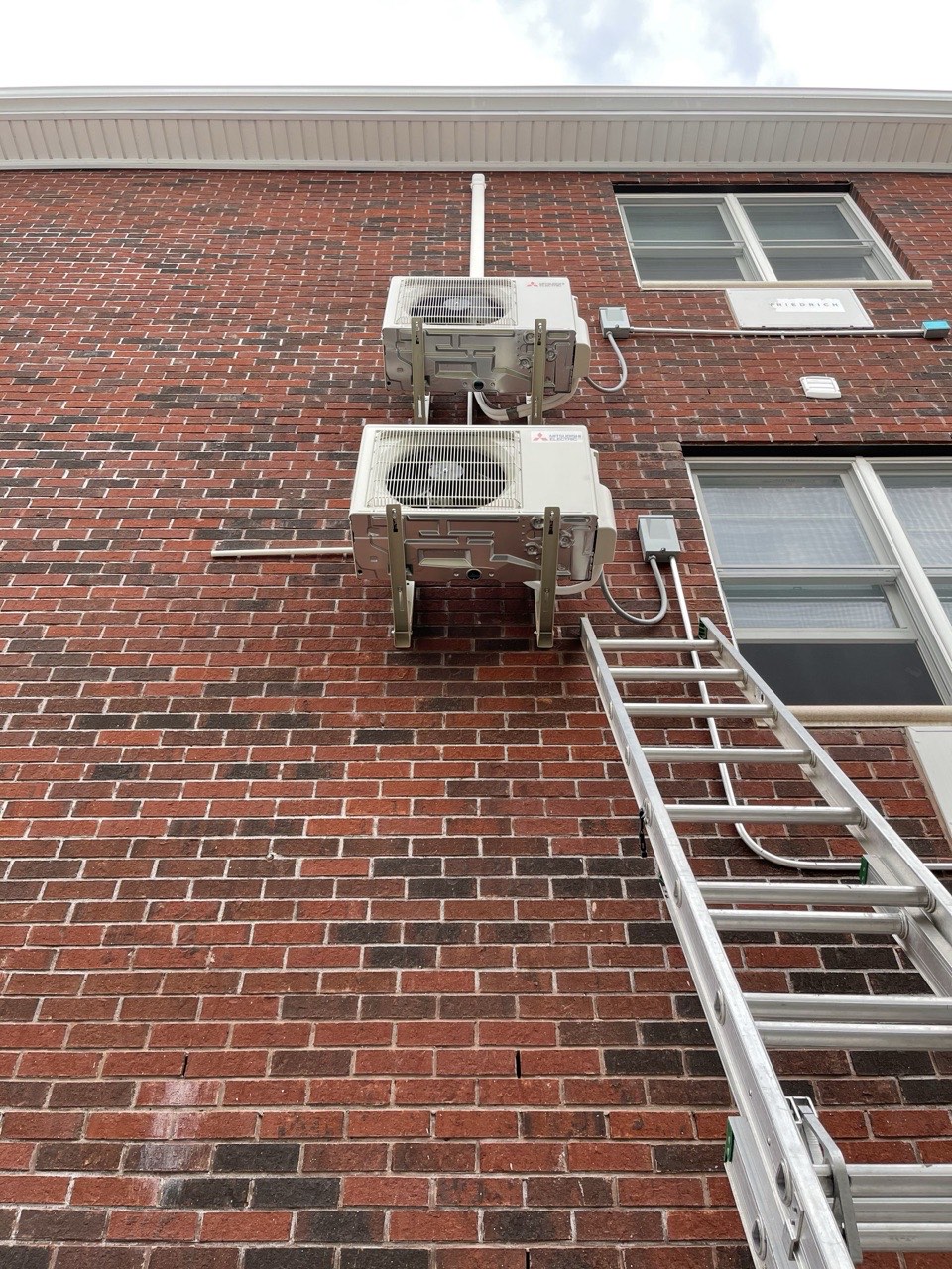 Two Mitsubishi Electric outdoor condensers wall-mounted on a Brooklyn brick building with installation ladder leaning against the facade