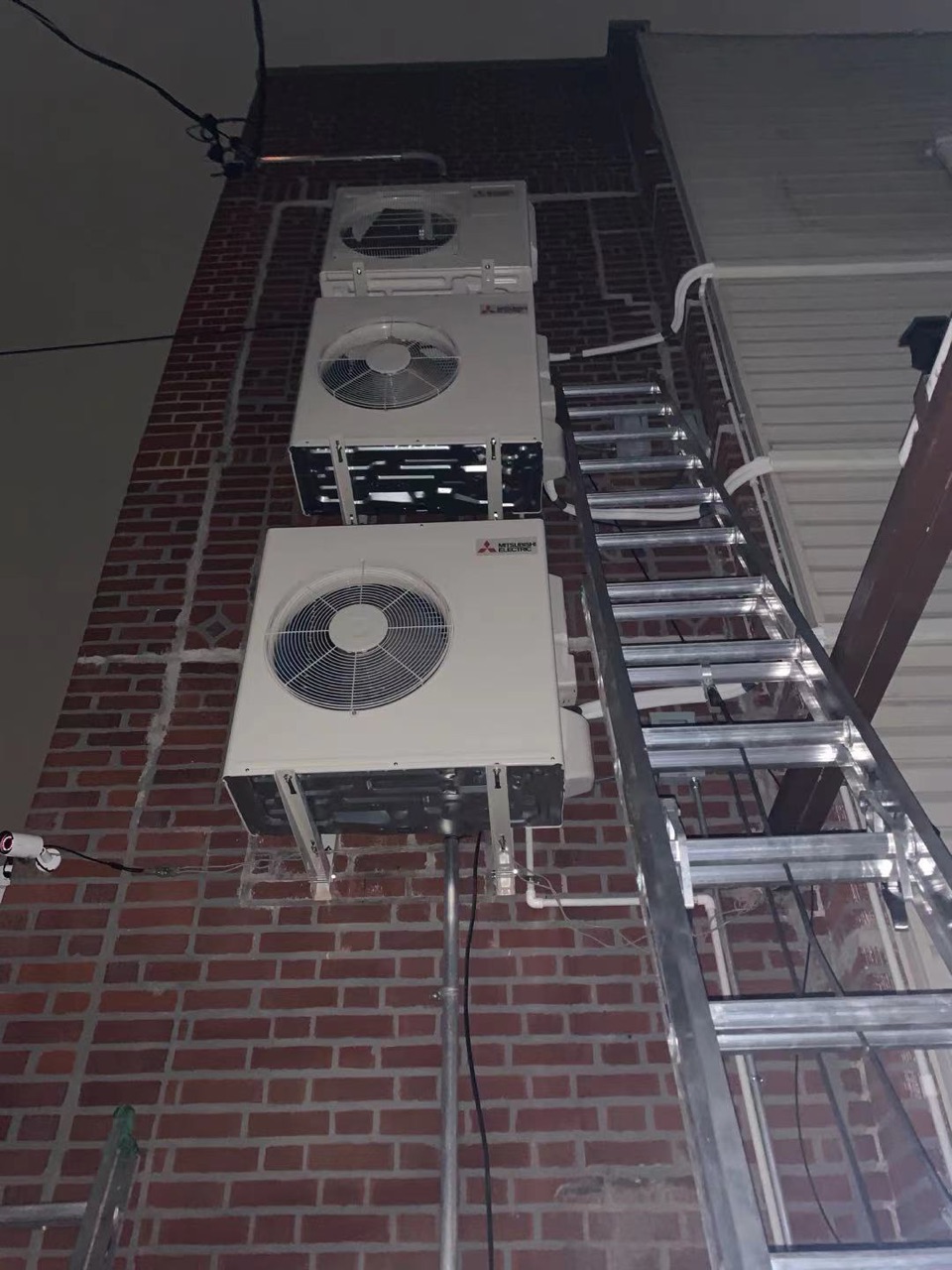 Three Mitsubishi Electric outdoor condensers stacked vertically on a brick wall illuminated at night