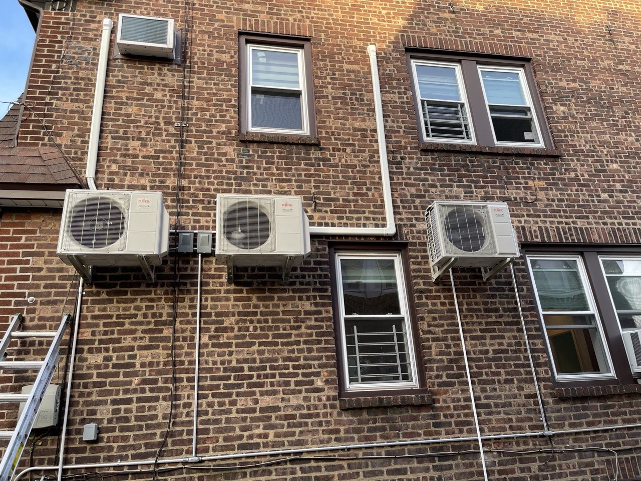 Multiple Fujitsu outdoor condenser units wall-mounted across the back of a Brooklyn brick building with window AC units alongside