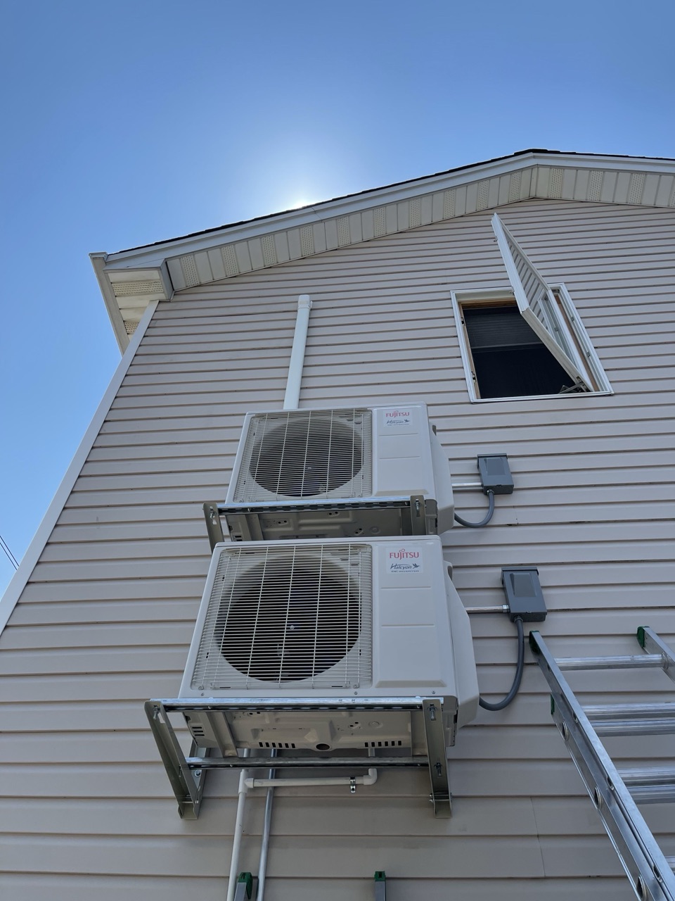 Two Fujitsu outdoor condenser units stacked on a wall-mount bracket against tan vinyl siding under a blue sky