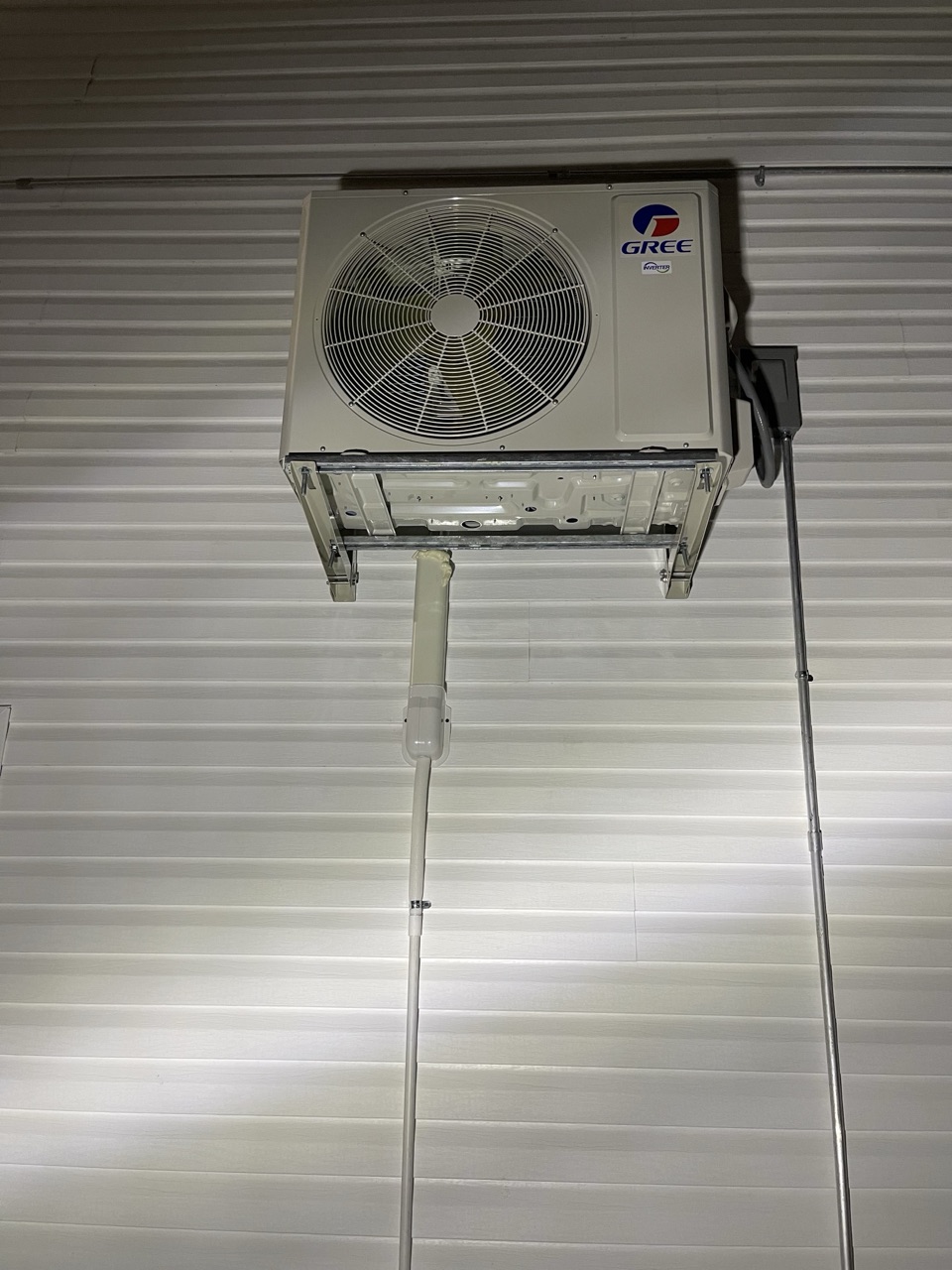 Gree outdoor condenser unit shown straight on, mounted on a metal-sided wall with neat drain line