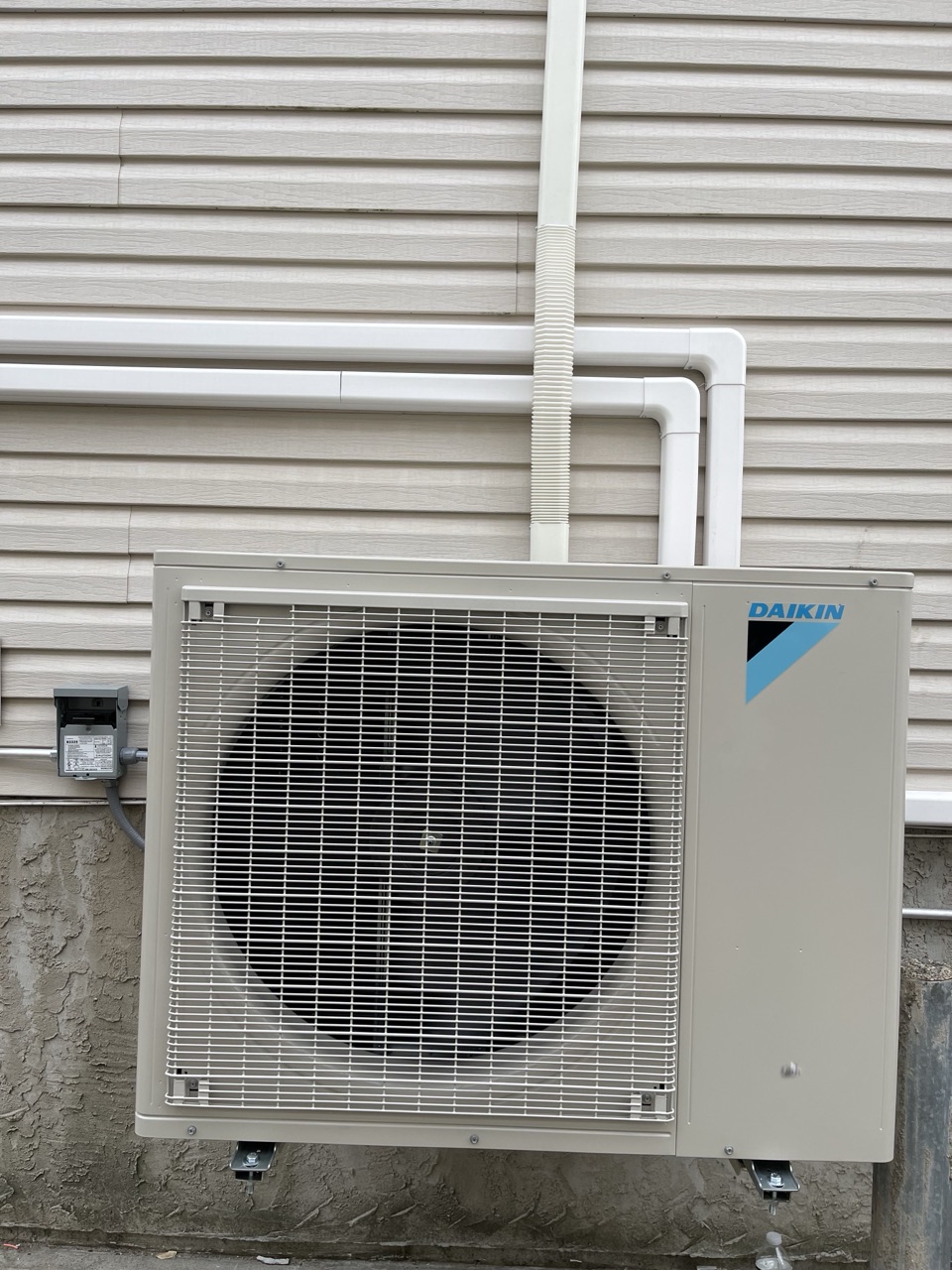 Daikin outdoor condenser unit installed at ground level against the tan-vinyl-sided exterior of a Brooklyn home